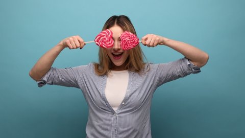 Portrait of satisfied young adult woman standing with two heart shape candies, looking at camera with toothy smile, wearing striped shirt. Indoor studio shot isolated on blue background.