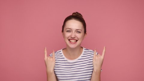 Yeah, that`s wonderful! Overjoyed woman showing rock and roll hand sign, screaming and gesturing to heavy metal, rock music, wearing striped T-shirt. Indoor studio shot isolated on pink background.