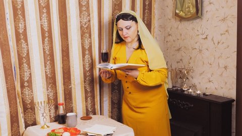 A Jewish woman in a wig and head covering makes a celebratory kdush with a glass in her hand and a siddur. Overall plan