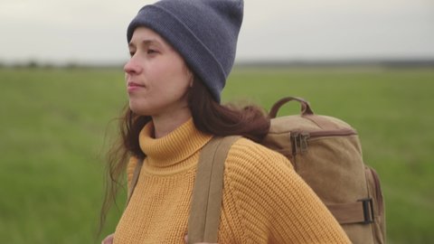 Young woman with long dark hair in orange sweater and beanie with backpack enjoys fresh air and windy weather hiking in field closeup