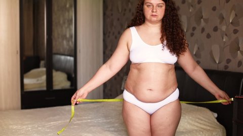 Curly overweight woman in white underwear stand near bed in bedroom, measure hips circumference with tape, commit and watch result. Fat burning treatment of thick cellulite body. Weight loss control.