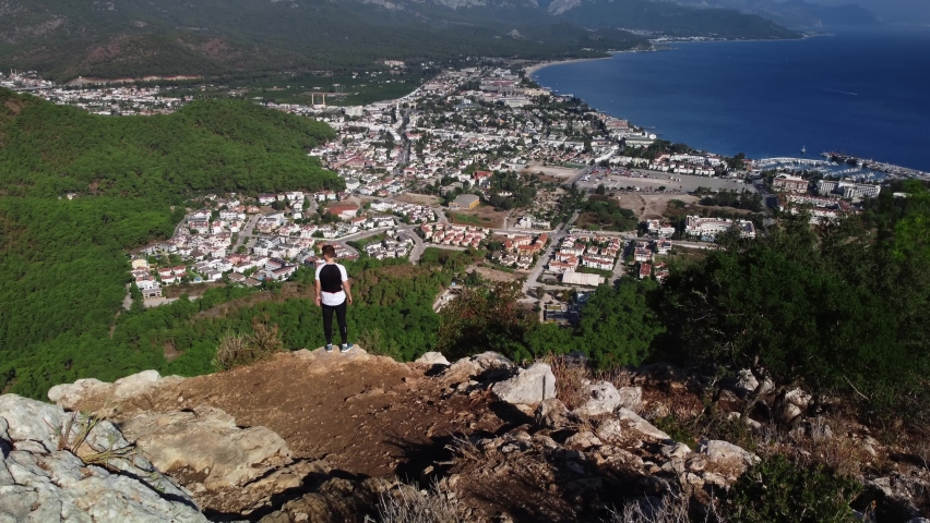 Caucasian man is standing on the edge of precipice enjoying the view of Kemer city and shoreline. Aerial footage of male tourist on peak of mountain looking at coastal town surrounded by green forest.