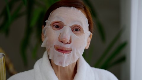 Headshot portrait of joyful mature woman drinking tasty vitamin smoothie smiling licking lips. Close-up front view of Caucasian retiree in moisturizing mask looking at camera posing at home in morning