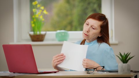 Mature Caucasian woman with menopause sitting at table using paper as hand fan. Portrait of tired redhead lady sweating working in office indoors. Female health concept