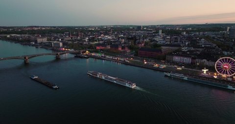 Drone aerial shot of Mainz two boats crossing in front of a summer wine festival at the Rhine river bank under orange sunset sky