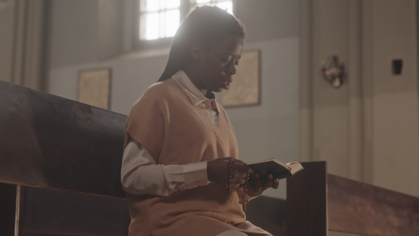 Medium slowmo of young African American female parishioner with rosary beads in hands reading Holy Bible and praying, sitting on wooden bench in Catholic church | Shutterstock HD Video #1089864039