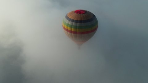 Beautiful colorful striped air balloon flying in cloudy sky atmosphere. Aerial cinematic clip. Thick fog of white moving clouds. Drone flight.
