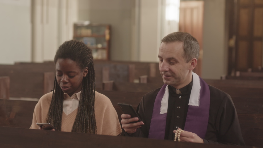 Slowmo of multiethnic priest and young female parishioner sitting next to each other on wooden bench in Catholic church using their smartphones | Shutterstock HD Video #1089864641