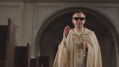 Tracking slowmo of confident young Caucasian pope wearing white vestment and black sunglasses walking along Catholic church with rosary beads in hands