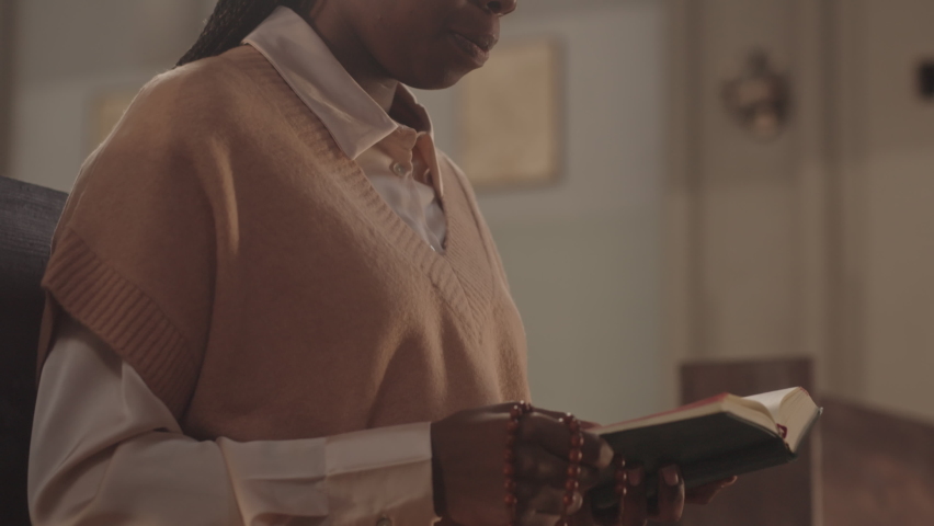 Slowmo of young Black woman holding rosary beads and Holy Bible in hands while praying to God sitting on wooden pew in Catholic church | Shutterstock HD Video #1089864813