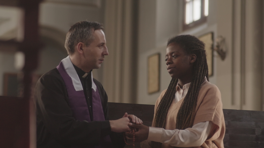 Medium shot of young African American woman talking to mature Caucasian priest and starting praying, sitting on wooden pew in beautiful Catholic church | Shutterstock HD Video #1089865139