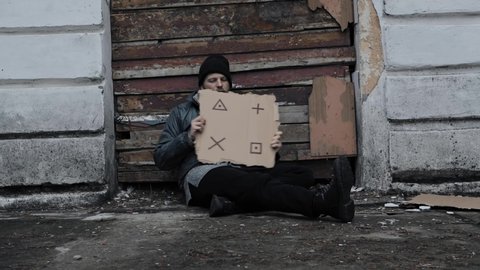 Homeless man sits holds piece of cardboard with markers for planar tracking. Immigrant. Below poverty line. Refugee looking for job. Tramp in dirty clothes, hat. Drunkenness. Bearded fugitive beggar