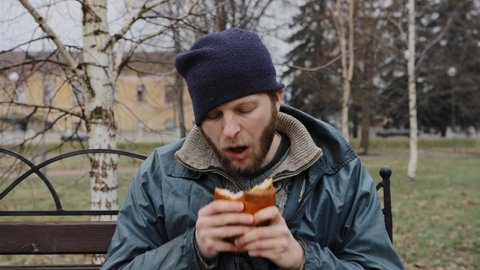 Hungry bearded homeless man sits on a bench and eats a bun in a city park. Below poverty line. Male tramp in dirty clothes with cap and hat. Beggar. Illegal immigrant. Drunkenness, alcoholism