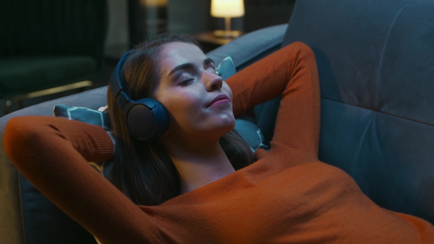 Young Woman is Lying at Home on the Couch with her Eyes Closed and Listening to Music on Headphones. Woman is Resting and Hearing to Online Radio. Relaxation, Meditation and Mindfulness. Royalty-Free Stock Footage #1089867993