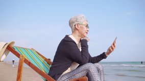 Asian elderly woman chatting with friends on video call while relaxing at the beach.