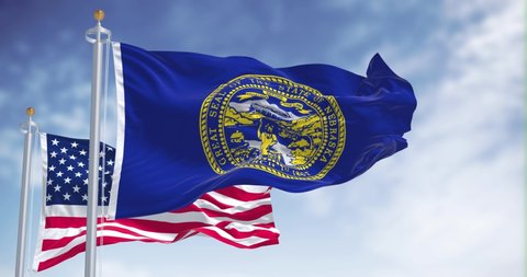 The Nebraska state flag waving along with the national flag of the United States of America. In the background there is a clear sky. Nebraska is a state in the Midwestern United States
