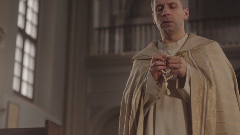 Medium slowmo of Caucasian pastor in white alb with rosary beads in hands walking along Catholic church praying to God