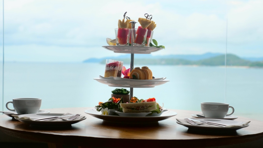 Traditional english high tea stand with selection of sweets, cakes, sandwiches, fruit canapes. Afternoon tea tray set with sweet treats on table. Tropical sea and mountains landscape background. Royalty-Free Stock Footage #1089874079