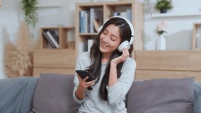 Positive Asian happy woman uses mobile phone and wears bluetooth headphones on couch. smile and feel good. Female listening MP3 music or watching a MV video on smartphone in the living room at home.
