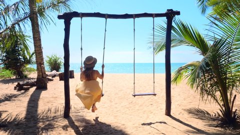Woman with hat enjoying freedom on swing in Phuket, Thailand. Life, adventure and travel concept. Happy young woman in long dress on swing enjoy summer vacation in tropical resort with sea view