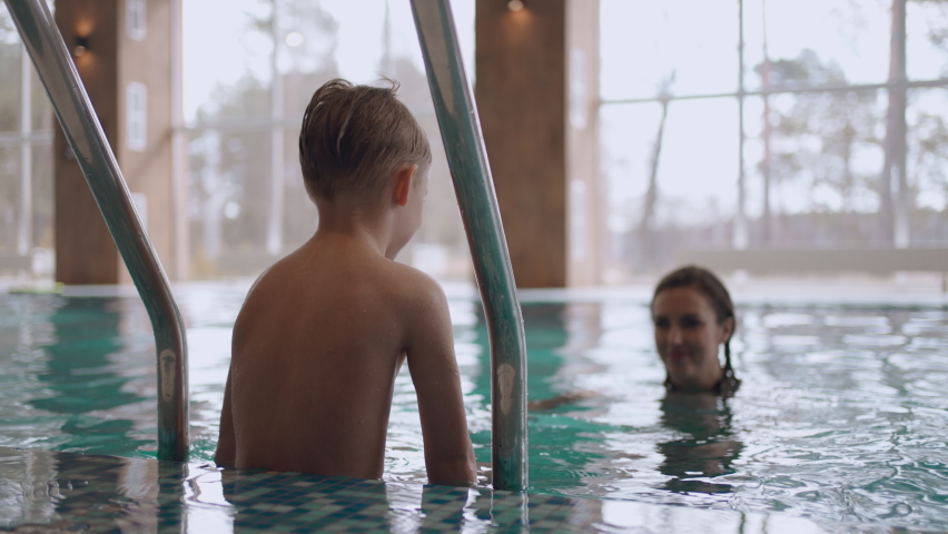 mother is teaching his son to swim in pool, joyful little boy is jumping in water to mom, happy family