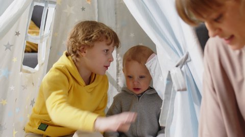 Adorable baby boy sitting in teepee tent and looking at mother while cheerful elder brother joining him