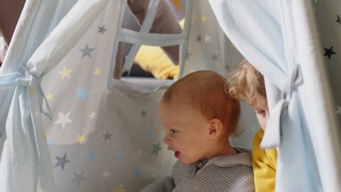 Cute baby boy sitting in teepee tent with elder brother and playing with joyful mother