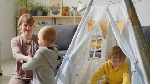 Little baby boy going to mother, taking her hand and walking with her in kids room while elder kid playing in teepee tent