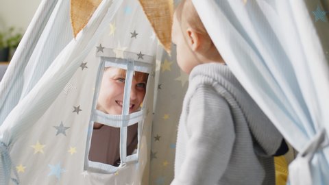 Cheerful little boy sitting with baby brother in teepee tent at home while joyous mother playing with them