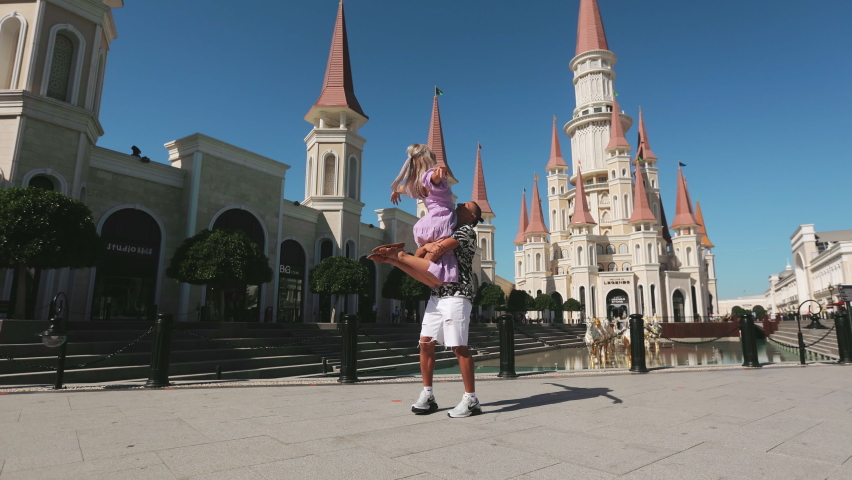 Happy handsome man holding on hands his charming woman and turning at theme park. Carefree caucasian couple dancing among famous resort at Belek.