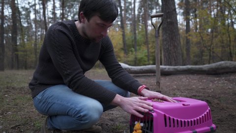 Upset male owner of dead pet says goodbye to animal before funeral in forest, sitting side by side with carrier and holding cat's favorite food in his hands. Theme is death of pet. Priceless moments.