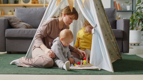 Mother sitting with baby and elder son in teepee tent at home and explaining kids how to play with toy