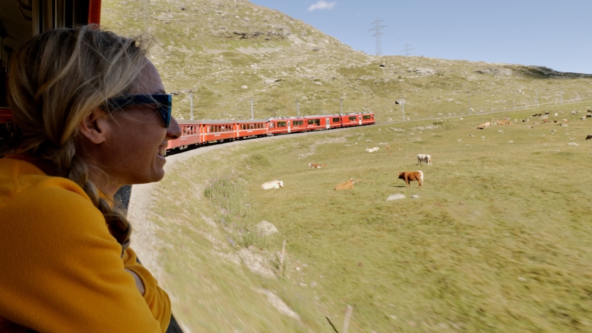 Woman traveller leaning out of train carriage window enjoys stunning scenery in the Alps. Female travelling Switzerland by train in summer 