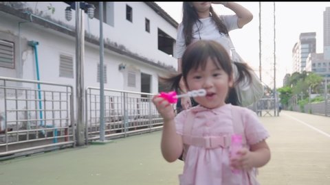 Playful asian girl blowing soap bubbles showing off playing with camera, asian child enjoy her time while walking back home from school, with small cute kid backpack, healthy fun preschooler age