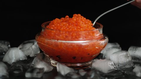 Tasting red salmon caviar salted roe. Hand with spoon takes of caviar from the glass jar. Bowls with red salmon salted roe caviar on tasting table on black background close-up slow motion