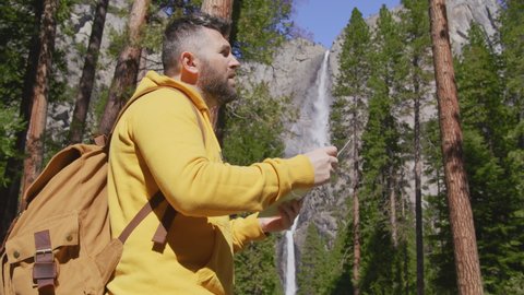 Contemporary Travel Guy with Tourist Backpack Standing in Nature Scenic, RED camera 6K. 30s Forest Hiker Traveler Man in Woods Hiking in Yosemite National park with Waterfall View, Tourism Concept.