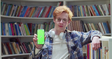 Portrait of young Caucasian red-haired male student in glasses smiling and showing smartphone with green screen and tracking motion in library. Chroma key. Guy with mobile phone demonstrating.