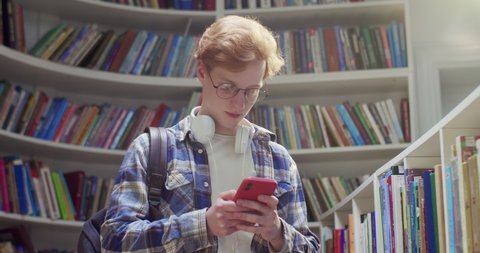 Young Caucasian red-haired male student in glasses and headphones chatting on smartphone and sending messages in library. Guy with red hair texting on mobile phone and tapping. Messages.