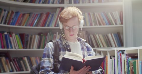 Caucasian red-haired male student in glasses and headphones flipping pages, reading book and searching for information in library. Guy with red hair with textbook at bookshelves listening to music.