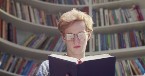 Close up of Caucasian red-haired young male student in glasses reading book and searching for information in library. Literature concept. Guy with red hair with textbook at bookshelves.