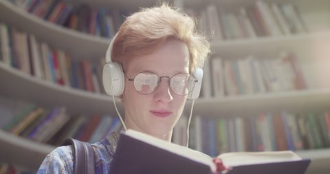 Close up of Caucasian red-haired male student in glasses and headphones flipping pages, reading book and searching for information in library. Guy with textbook at bookshelves listening to music.