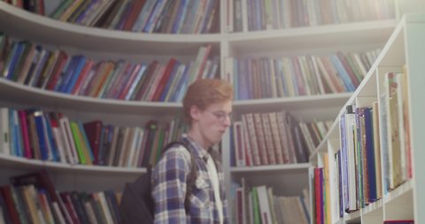Young red-haired Caucasian male student with backpack walking in library, choosing textbook to read. Taking out book from shelf. Literature concept. Guy coming to bibliotheca to study.