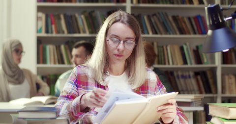 Portrait shot of young blond joyful Caucasian female student in glasses reading book in library, then rising head and smiling to camera. Pretty happy woman with textbook in hands. Learning concept.