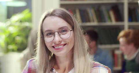 Close up of pretty joyful young blonde Caucasian female student in glasses looking at camera and smiling. Indoor. Beautiful woman studying in library. Portrait shot.