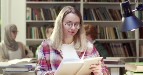 Portrait of young blonde cheerful Caucasian female student in glasses reading book in library, then rising head and smiling to camera. Pretty woman with textbook in hands. Educating concept.