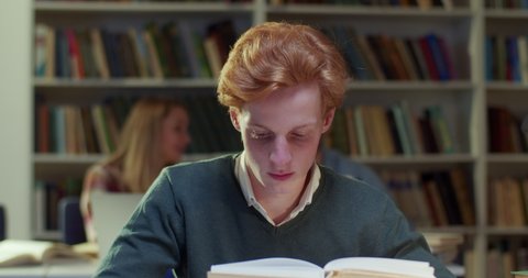 Close up of clever Caucasian young handsome red-haired guy reading textbook in library and flipping pages. Smart male student with red hair studying with book in cozy bibliotheca.