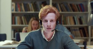 Close up of red-haired Caucasian male in headset videochatting for education vlogand talking to camera in library. Young man speaking, having videochat. Video call. Tutoring. Educational vlogging.