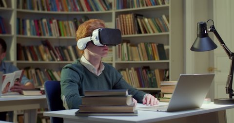 Caucasian young male student in VR headset sitting in library space, scrolling and moving hands in air and having virtual reality experience. Metaverse studying. Study in virtuality concept.