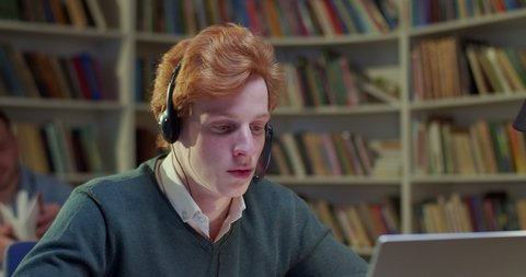 Close up of Caucasian red-haired male in headset talking and videochatting on laptop in library. Young man speaking and having videochat online via webcam. Video call. Tutoring.