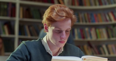 Close up of Caucasian young handsome red-haired guy reading textbook in library and flipping pages. Male student with red hair studying with book in cozy bibliotheca.